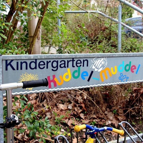 Zu sehen ist der Schriftzug "Kindergarten Kuddelmuddel", der auf einem Fahrradständer zu lesen ist. Eine kleine Maus ist ebenfalls abgebildet. Im Hintergrund liegt viel Laub und Grün. Die Lenker eines City-Rollers und eines kleinen Fahrrads sind ebenfal